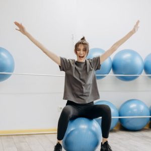 A cheerful woman in active wear exercises with a yoga ball indoors, promoting wellness.